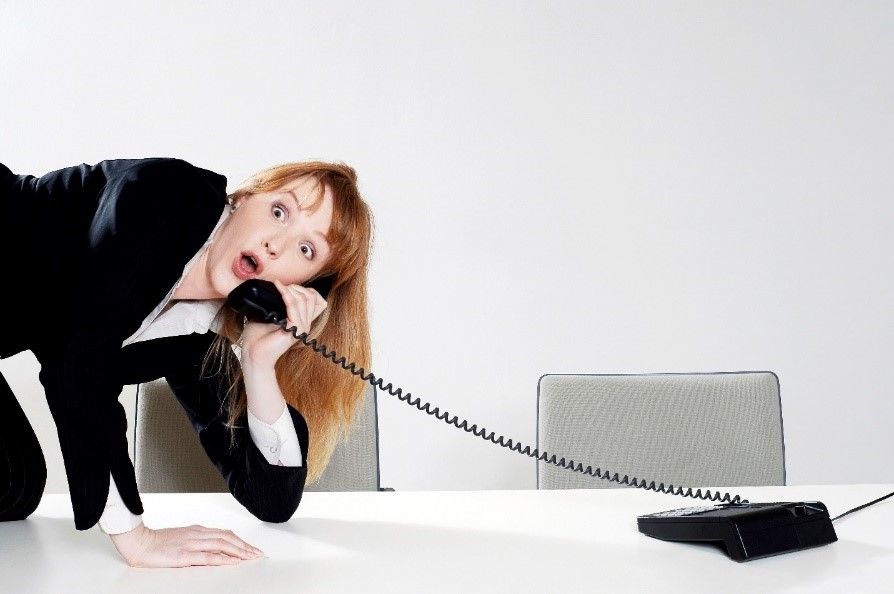 A dark-blonde woman in a black suite is on top of the desk answering a phone call. She has a look of surprise on her face.