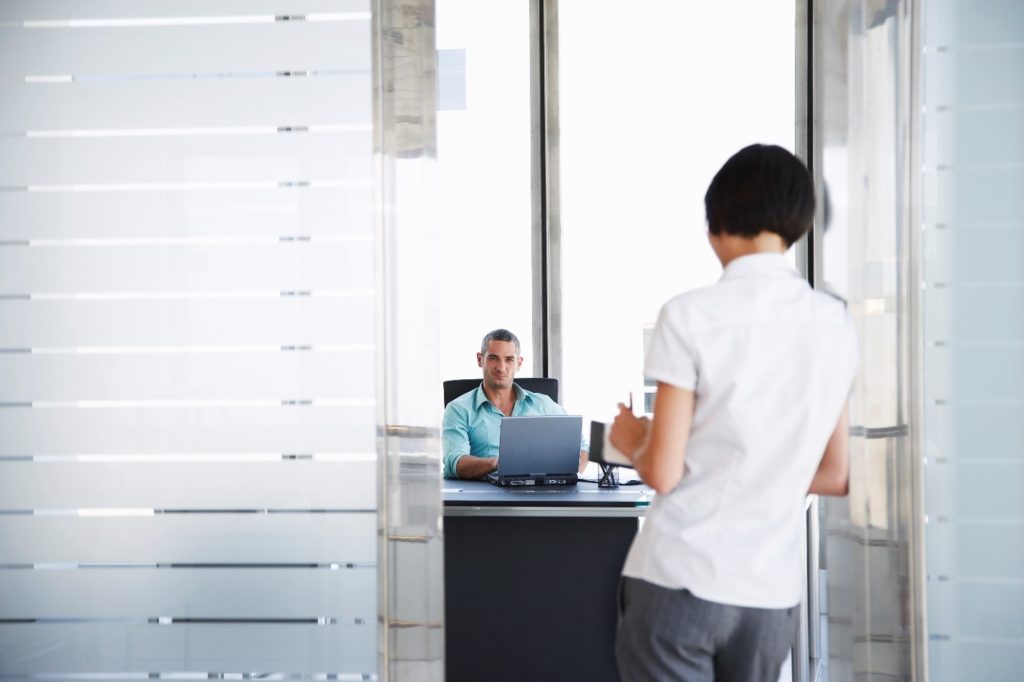 A person wearing a white shirt and grey pants is leaning against an open door to a man's office. We only see the back of the leaner. The man in the office is sitting at his desk working on a laptop, looking toward the door. He's wearing a light blue shirt.
