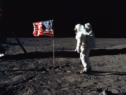 Man in white space suite standing on the moon by an American flag.