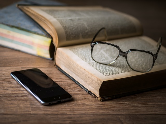Eyeglasses sitting on open book on table next to mobile phone.